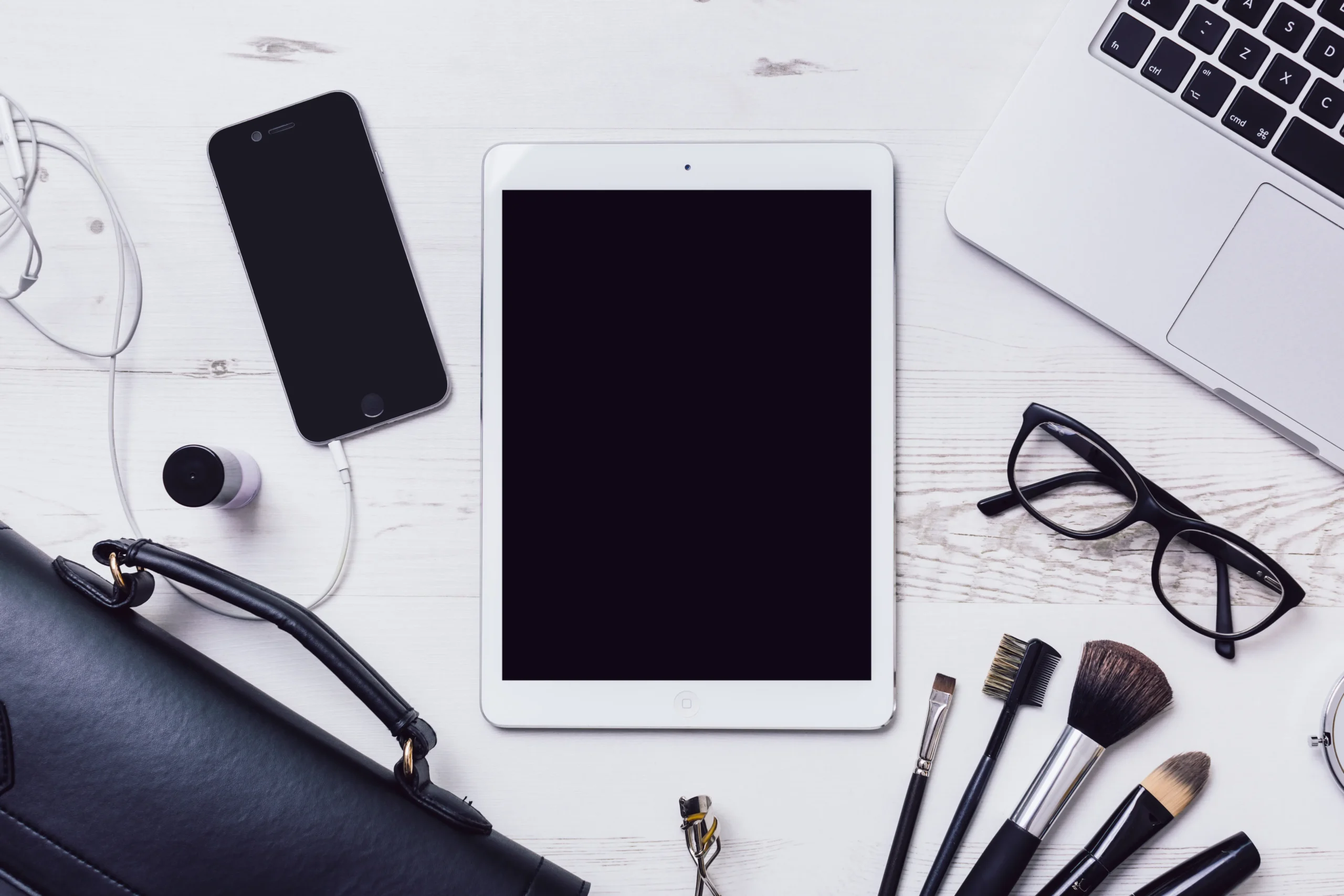 Flat lay of a phone, tablet, earbuds, and makeup brushes on a desk