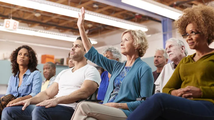 Diverse crowd at a public meeting with one person raising a hand