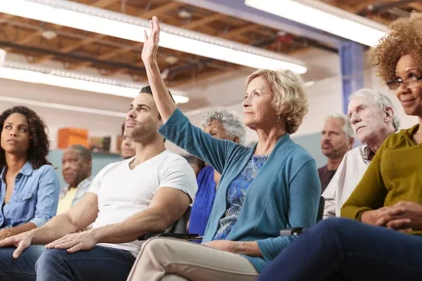 Diverse crowd at a public meeting with one person raising a hand